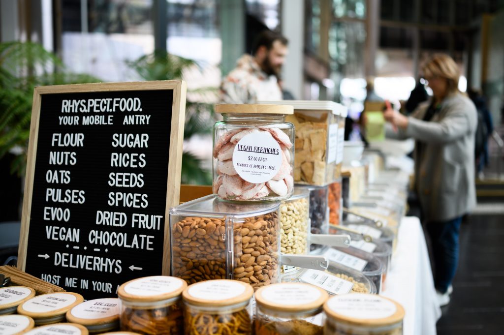 A stallholder engages a customer with their own bag in the background amongst the clear containers of food (vegan meringes, nuts, crackers, pretzels) with round labels stacked and organised with scoopers on the table and a sign describing the mobile pantry - flour, nutes, oats, pulses, evoo, vegan, sugar, rices, seeds, spices, dried fruit, chocolate.