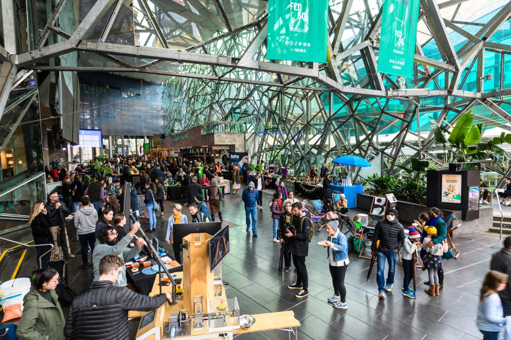 A busy atrium at Fed Sq with stalls, people walking around and the Precious Plastic group working machinary with people watching. 