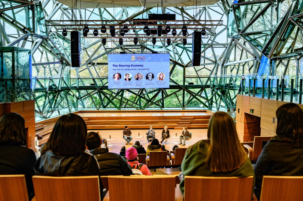 An amplitheatre full of people looking down towards a panel of speakers with a giant screen reading 'The Sharing Economy'.