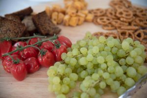 (From left to right) cherry tomatoes on the vine, banana cake, pretzels, apricot bites, and grapes sitting on a wooden board.